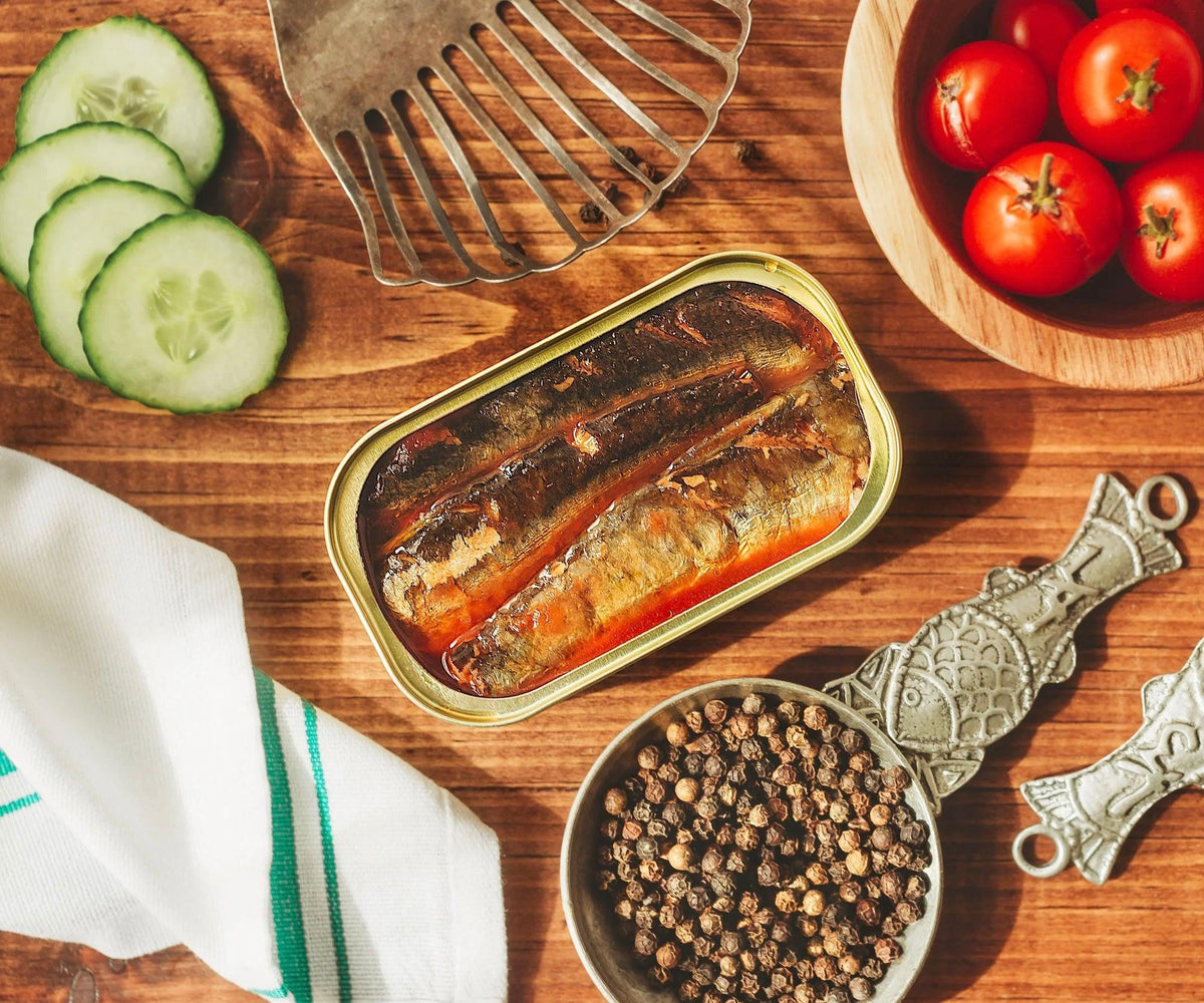 Open tin of sardines in escabeche sauce on a wooden board, with sliced cucumber, cherry tomatoes and peppercorns.